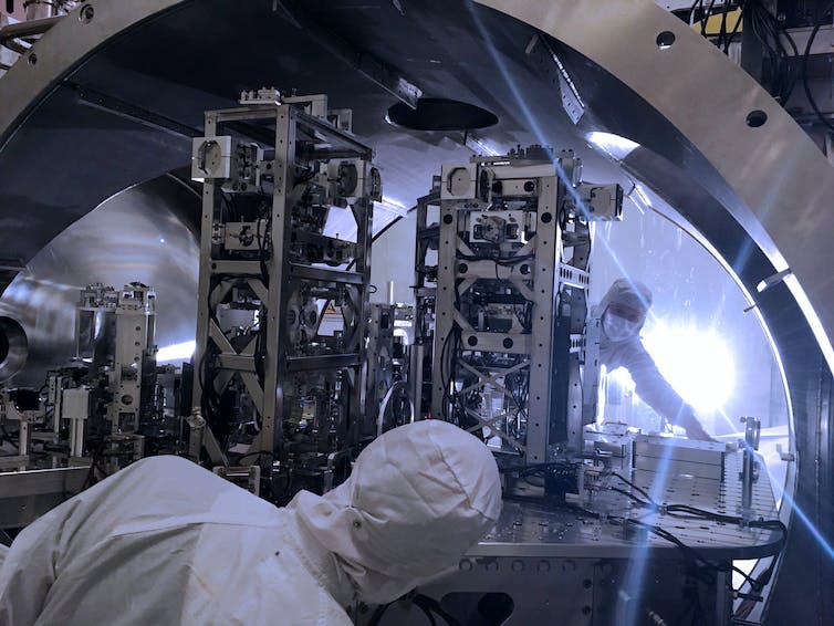 Two people in white lab outfits working on complicated machinery.